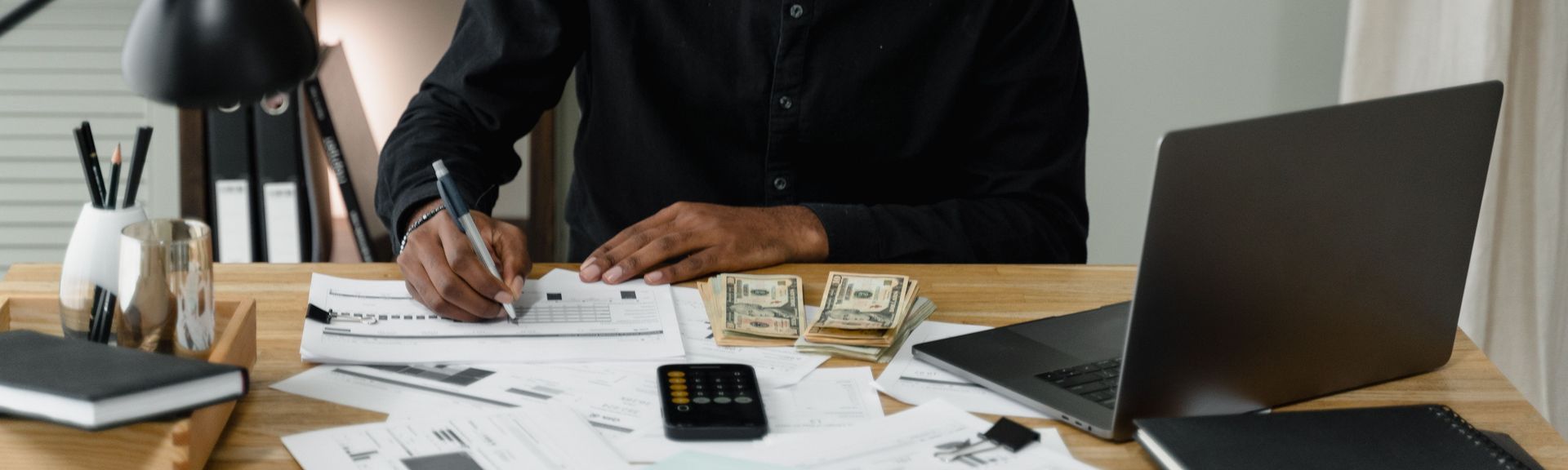 An adult man calculates expenses, using a laptop and documents at a desk in a home office setting.